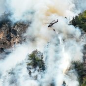 Ein Löschhubschrauber fliegt durch dichten Rauch über einer felsigen Waldschlucht während eines Waldbrandes in Kanada. Kanada-Rundreise
