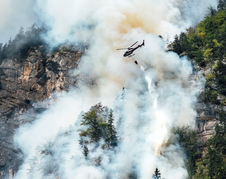 Ein Löschhubschrauber fliegt durch dichten Rauch über einer felsigen Waldschlucht während eines Waldbrandes in Kanada. Kanada-Rundreise