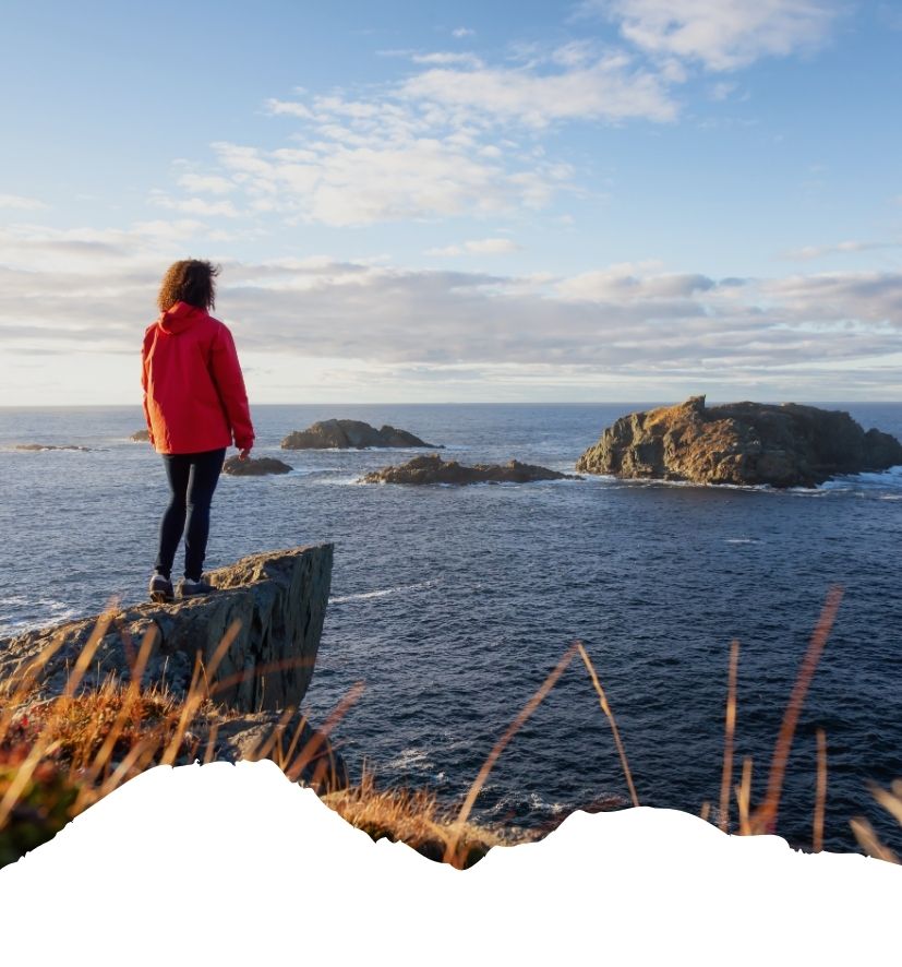 Frau in roter Jacke steht auf einem Felsen und blickt auf das Meer mit Inseln im Hintergrund.