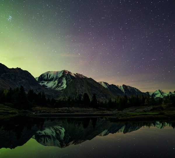 Nordlichter und Sterne über einer Berglandschaft mit ruhigem Wasser, das die Berge reflektiert.