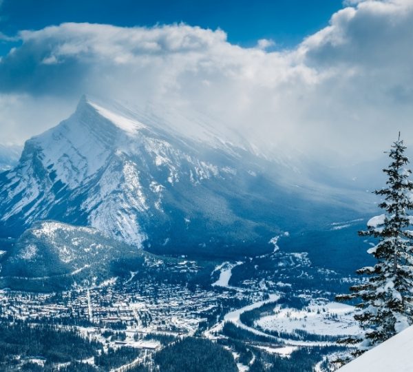 Schneebedeckter Berggipfel mit Wolken und einem Baum im Vordergrund in einer Berglandschaft.