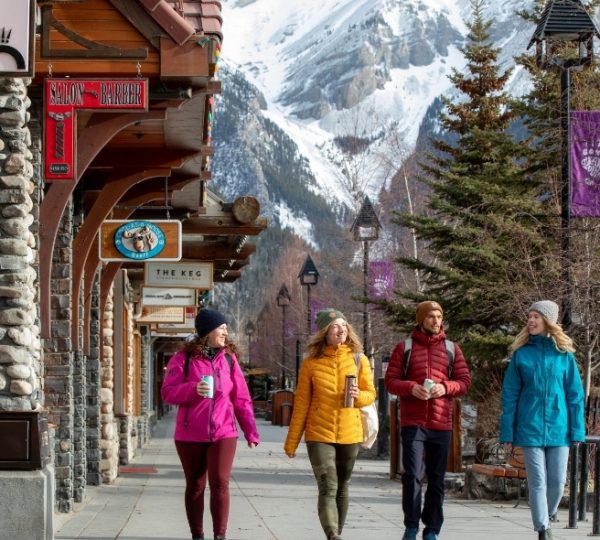 Vier Personen in Winterkleidung gehen auf einem Gehweg in Banff, umgeben von Geschäften und Bergen.