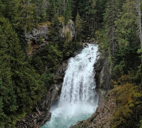 Ein Wasserfall fließt über Felsen in einem bewaldeten Gebiet mit grünen Bäumen.