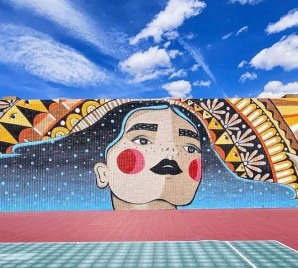 Wandmalerei einer Frau mit langen Haaren und buntem, geometrischem Hintergrund auf einer Mauer.