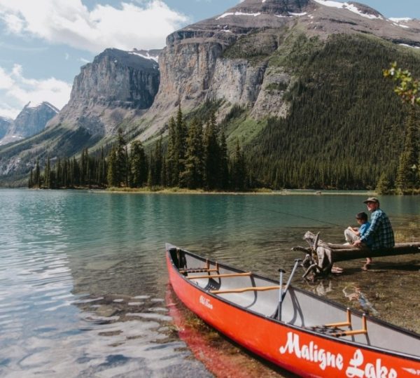 Ein rotes Kanu am Ufer des Maligne Lake mit einem Angler und einem Kind im Hintergrund.