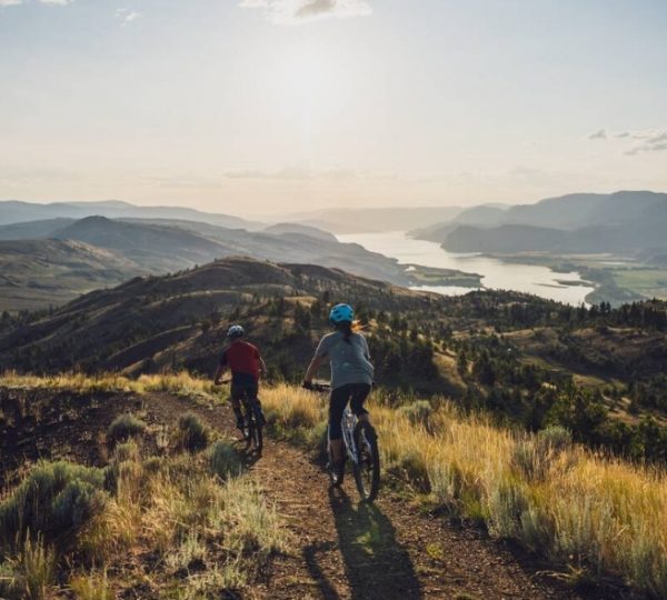 Zwei Mountainbiker fahren auf einem Pfad mit Blick auf eine Landschaft und einen Fluss im Hintergrund.
