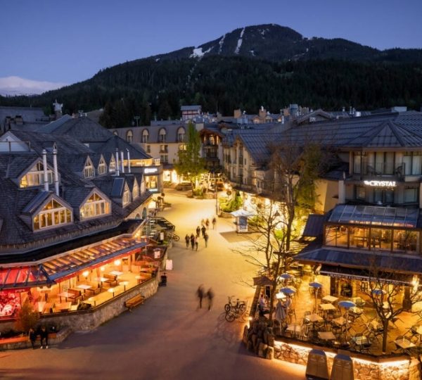 Abendliche Ansicht des Dorfes Whistler mit beleuchteten Gebäuden und einem Berg im Hintergrund.