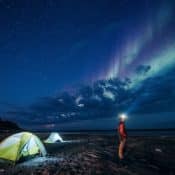Ein Mann mit Stirnlampe steht neben zwei Zelten unter einem sternenklaren Himmel mit Nordlichtern.