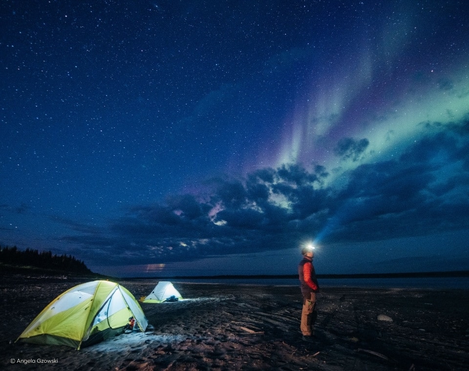 Ein Mann mit Stirnlampe steht neben zwei Zelten unter einem sternenklaren Himmel mit Nordlichtern.
