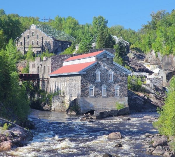 Historisches Gebäude La Pulperie am Ufer eines Flusses in Chicoutimi, umgeben von Bäumen.