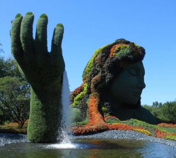 Große Skulptur einer Frau mit Pflanzen und Blumen, die eine Hand hebt, vor einem Wasserfall.