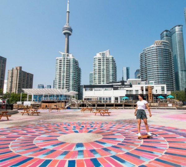 Eine Frau fährt Skateboard auf einem bunten Boden mit Wolkenkratzern und dem CN Tower im Hintergrund in Toronto.