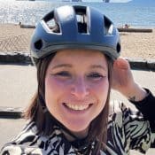 Frau mit kurzem Haar und Fahrradhelm lächelt in die Kamera am Strand.