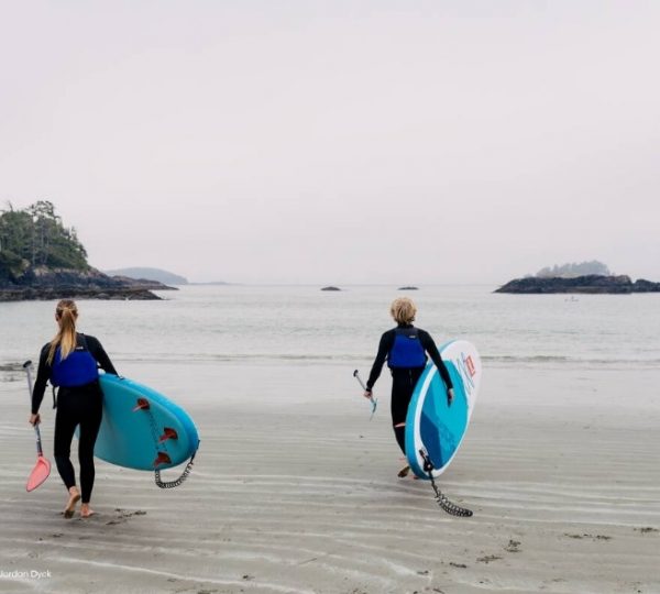 Zwei Personen tragen Stand-Up-Paddle-Boards am Strand entlang in Richtung Wasser.