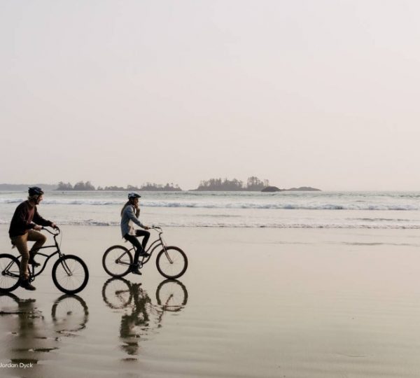 Zwei Radfahrer fahren am Strand entlang, während die Wellen im Hintergrund brechen.