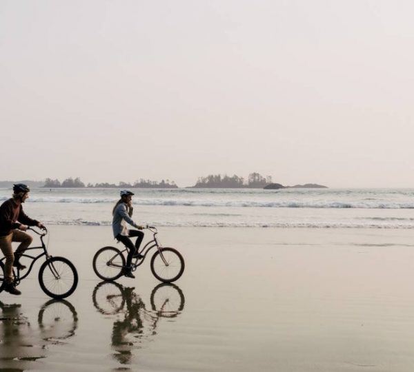Zwei Personen fahren auf Fahrrädern entlang eines Strandes mit Wasser im Hintergrund. Kanada-Rundreise