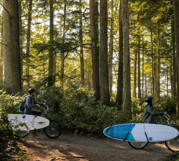 Zwei Personen mit Surfbrettern auf Fahrrädern in einem Waldgebiet mit hohen Bäumen. Kanada-Rundreise
