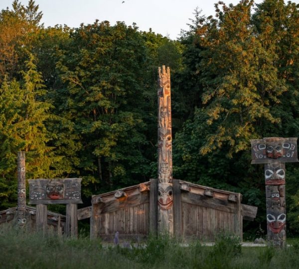 Totempfähle mit traditionellen Schnitzereien und eine Holzstruktur in einer bewaldeten Umgebung. Kanada-Rundreise