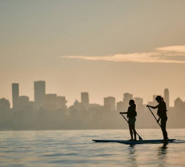 Zwei Personen paddeln auf Stand-Up-Paddle-Boards vor einer silhouettierten Stadtlandschaft bei Sonnenuntergang. Kanada-Rundreise