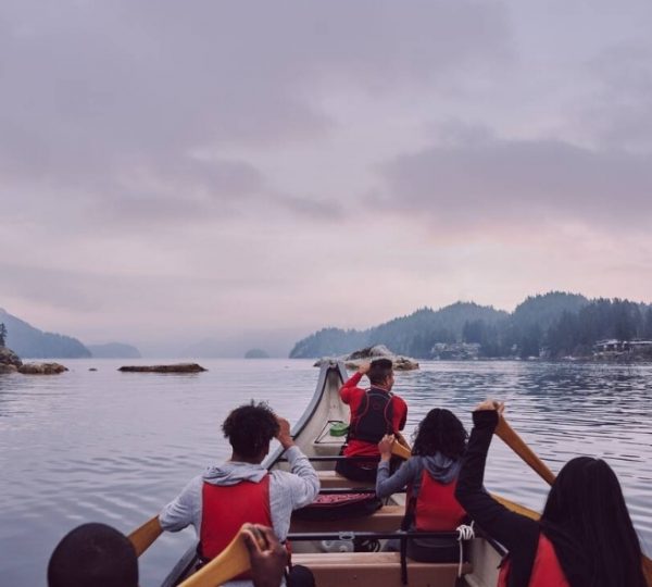 Eine Gruppe von Personen paddelt in einem Kanu auf ruhigem Wasser mit bewaldeten Ufern im Hintergrund. Kanada-Rundreise