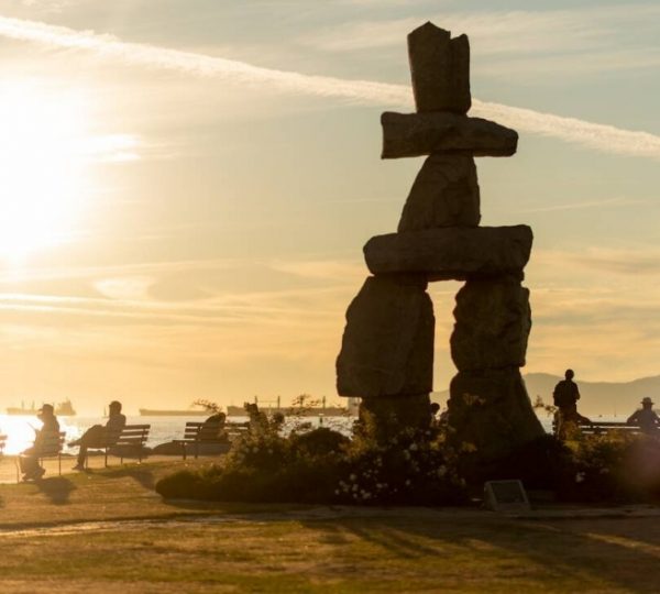 Inukshuk-Skulptur am Strand mit Menschen im Hintergrund während des Sonnenuntergangs. Kanada-Rundreise
