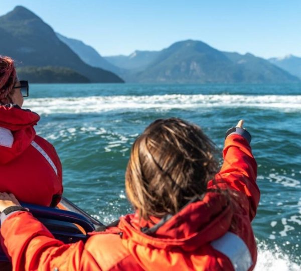 Zwei Personen in roten Jacken sitzen auf einem Boot und zeigen auf das Wasser und die Berge im Hintergrund. Kanada-Rundreise