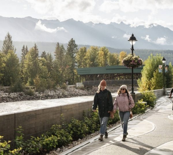 Ein Paar geht entlang eines Flusses mit Bergen im Hintergrund und einer Fahrradfahrerin auf dem Gehweg.
