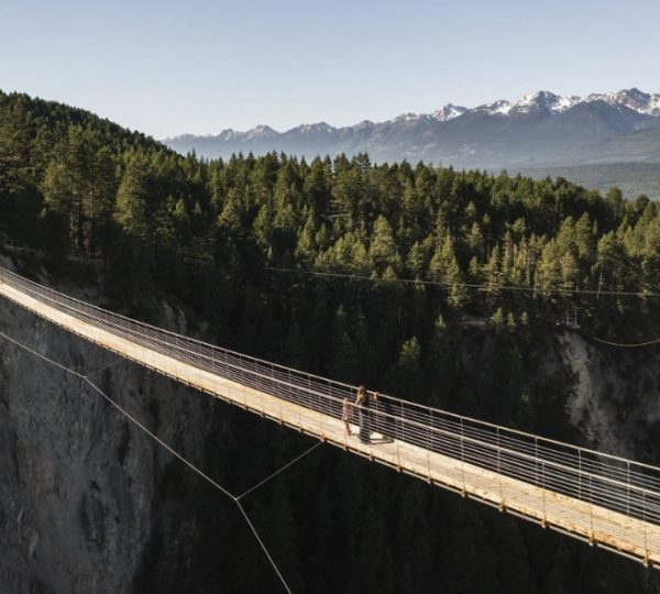 Eine Person überquert eine hängende Brücke in den kanadischen Rocky Mountains mit bewaldeten Hügeln im Hintergrund. Kanada-Rundreise
