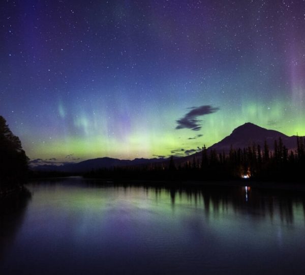 Nordlichter leuchten über einem ruhigen Fluss mit Bergen im Hintergrund und einem klaren Nachthimmel.