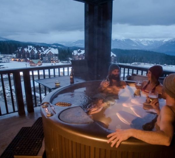 Drei Personen sitzen in einem Whirlpool auf einer Terrasse mit Blick auf die Berge und den Schnee. Kanada-Rundreise