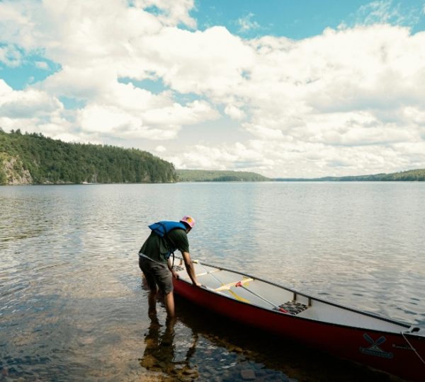 Eine Person, die ein rotes Kanu am Ufer eines ruhigen Gewässers vorbereitet. Kanada-Rundreise