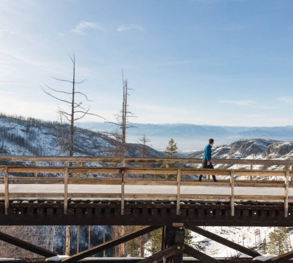 Eine Person geht auf einer Holzbrücke in einer schneebedeckten Landschaft mit Bergen im Hintergrund.