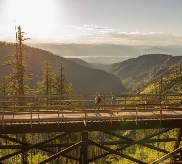 Zwei Personen gehen über eine Holzbrücke in einer Berglandschaft mit Bäumen und Bergen im Hintergrund.