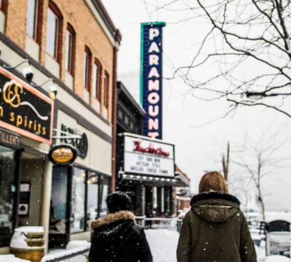 Zwei Personen gehen in winterlicher Kleidung auf einer schneebedeckten Straße mit Geschäften und einem Kino. Kanada-Rundreise