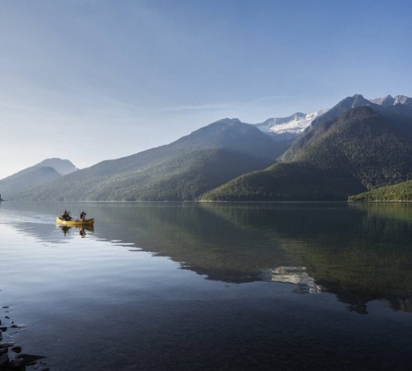 Zwei Kajakfahrer auf einem ruhigen See, umgeben von Bergen und klarem Himmel. Kanada-Rundreise