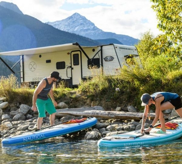 Zwei Personen bereiten ihre Stand-Up-Paddle-Boards am Ufer eines Gewässers vor, im Hintergrund ein Wohnmobil.