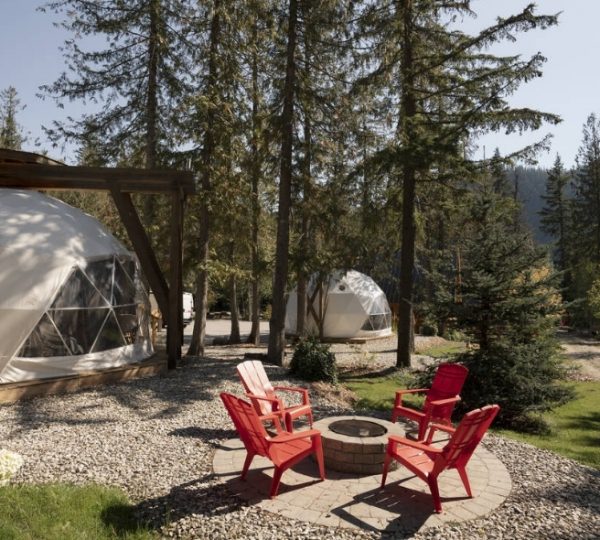 Zwei weiße Glamping-Dome mit roten Stühlen und einem Feuerplatz in einem Waldgebiet. Kanada-Rundreise