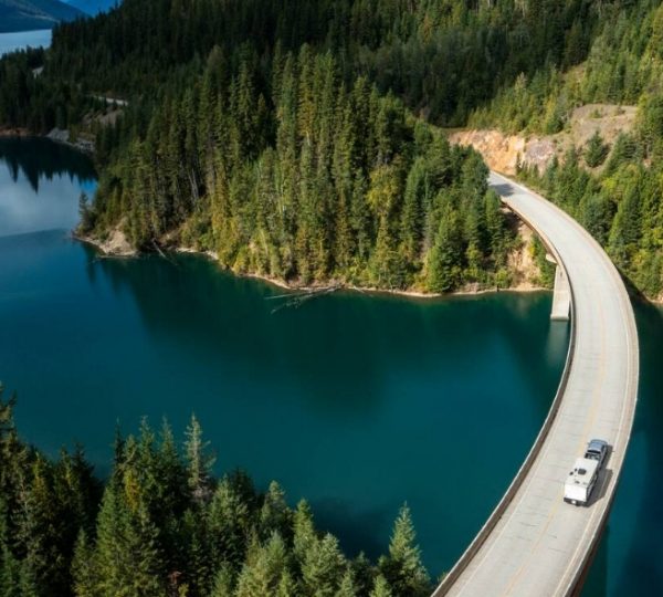 Eine kurvenreiche Straße führt entlang eines klaren Gewässers in Westkanada, umgeben von Bäumen.