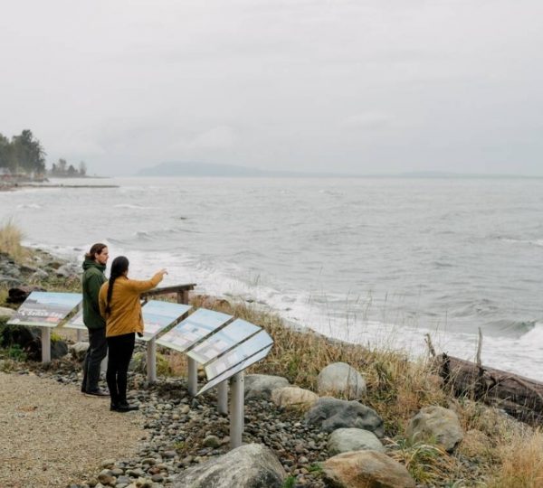 Zwei Personen stehen am Strand und betrachten Informationstafeln über die Küstenlandschaft von Parksville. Kanada-Rundreise