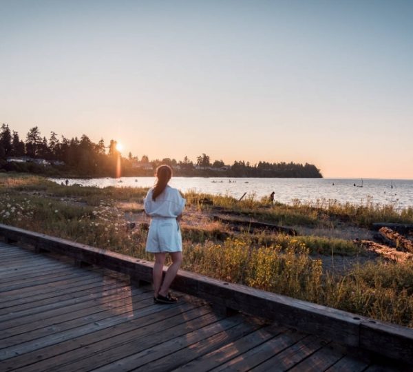 Frau in weißem Kleid steht am Strand und blickt auf den Sonnenuntergang über dem Wasser. Kanada-Rundreise