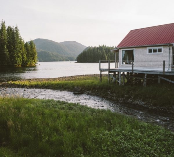 Ein Holzhaus mit rotem Dach am Ufer eines Gewässers in Westkanada, umgeben von Bäumen und Bergen. Kanada-Rundreise