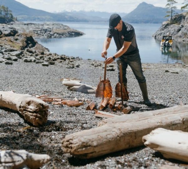 Mann, der Holzstücke am Strand aufnimmt, mit Wasser im Hintergrund und Bergen in der Ferne. Kanada-Rundreise