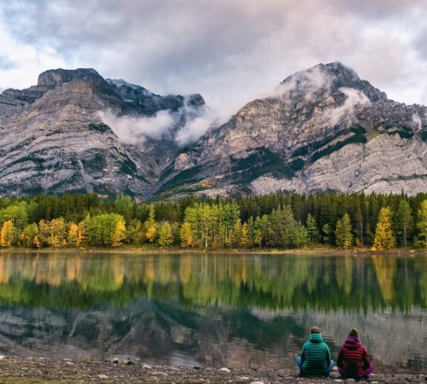 Zwei Personen sitzen am Ufer eines ruhigen Sees mit Bergen im Hintergrund und reflektierenden Wolken. Kanada-Rundreise