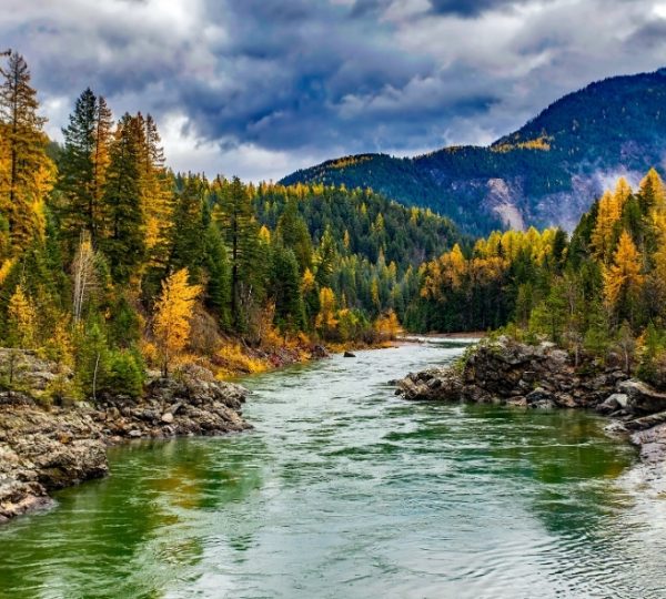 Ein Fluss fließt durch eine herbstliche Landschaft mit Bäumen und Bergen im Hintergrund. Kanada-Rundreise