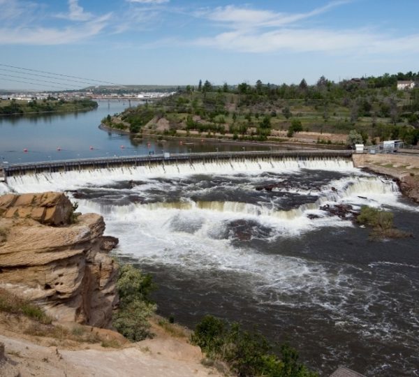 Wasserfall, der über einen Staudamm fließt, umgeben von Wasser und Vegetation. Kanada-Rundreise