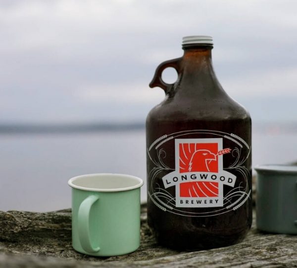 Ein Longwood Brewery Growler mit zwei Tassen auf einem Holzstamm am Wasser. Kanada-Rundreise