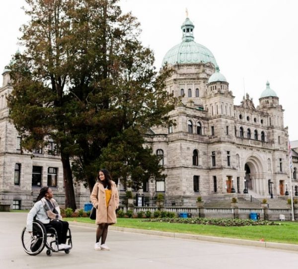 Zwei Frauen, eine im Rollstuhl, stehen vor einem historischen Gebäude in Victoria, Kanada. Kanada-Rundreise