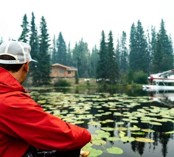 Eine Person in roter Jacke sitzt am Wasser, umgeben von Seerosen und Bäumen. Kanada-Rundreise