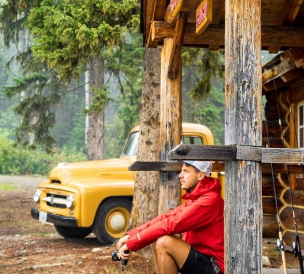 Mann in roter Jacke sitzt auf einer Holzveranda vor einer Hütte mit einem gelben Oldtimer im Hintergrund. Kanada-Rundreise