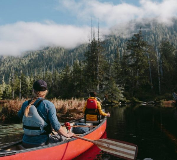 Zwei Personen paddeln in einem roten Kanu auf einem ruhigen Gewässer umgeben von Bäumen und Bergen. Kanada-Rundreise